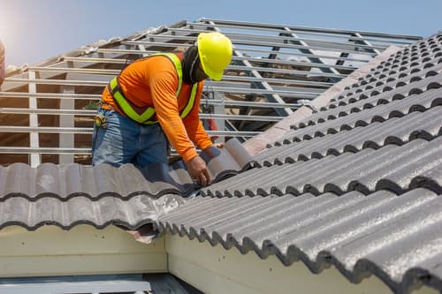 A roofer wearing protective gear replaces tiles on a house roof.